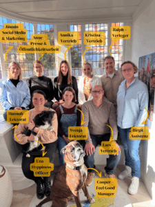 Teamfoto des CalmeMara Verlag. Sie sitzen in zwei Reihen voreinander und lächeln in die Kamera. Außerdem sind die beiden Verlagshunde zu sehen.